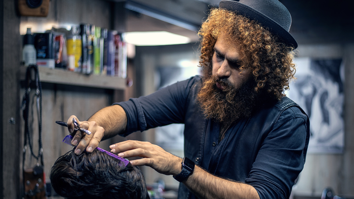 A curly-haired barber cuts his client's hair as they discuss digital marketing strategy.